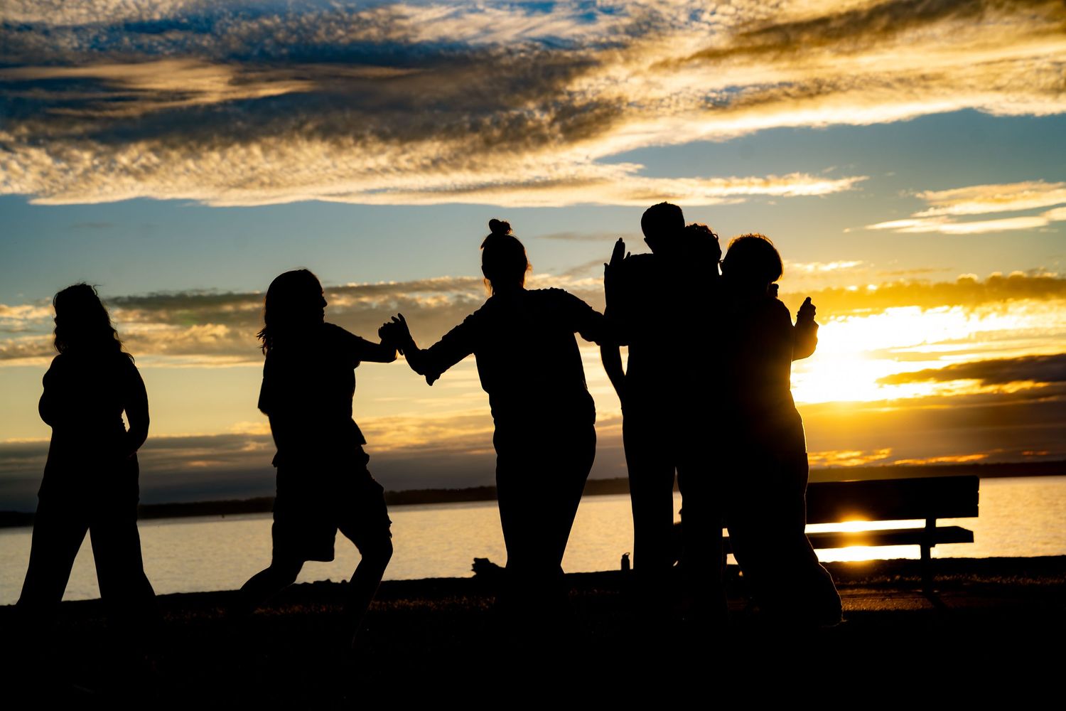 Silhouettes of friends holding hands and embracing at sunset by the water with dramatic cloudy sky.