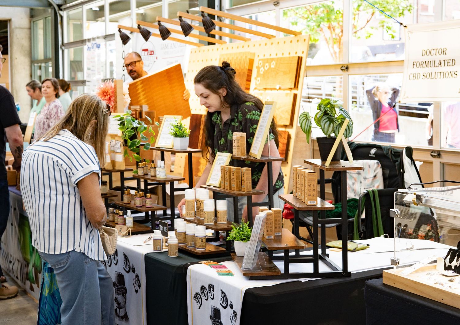 Shoppers browse handmade wooden crafts and plants at an indoor artisan market stall.