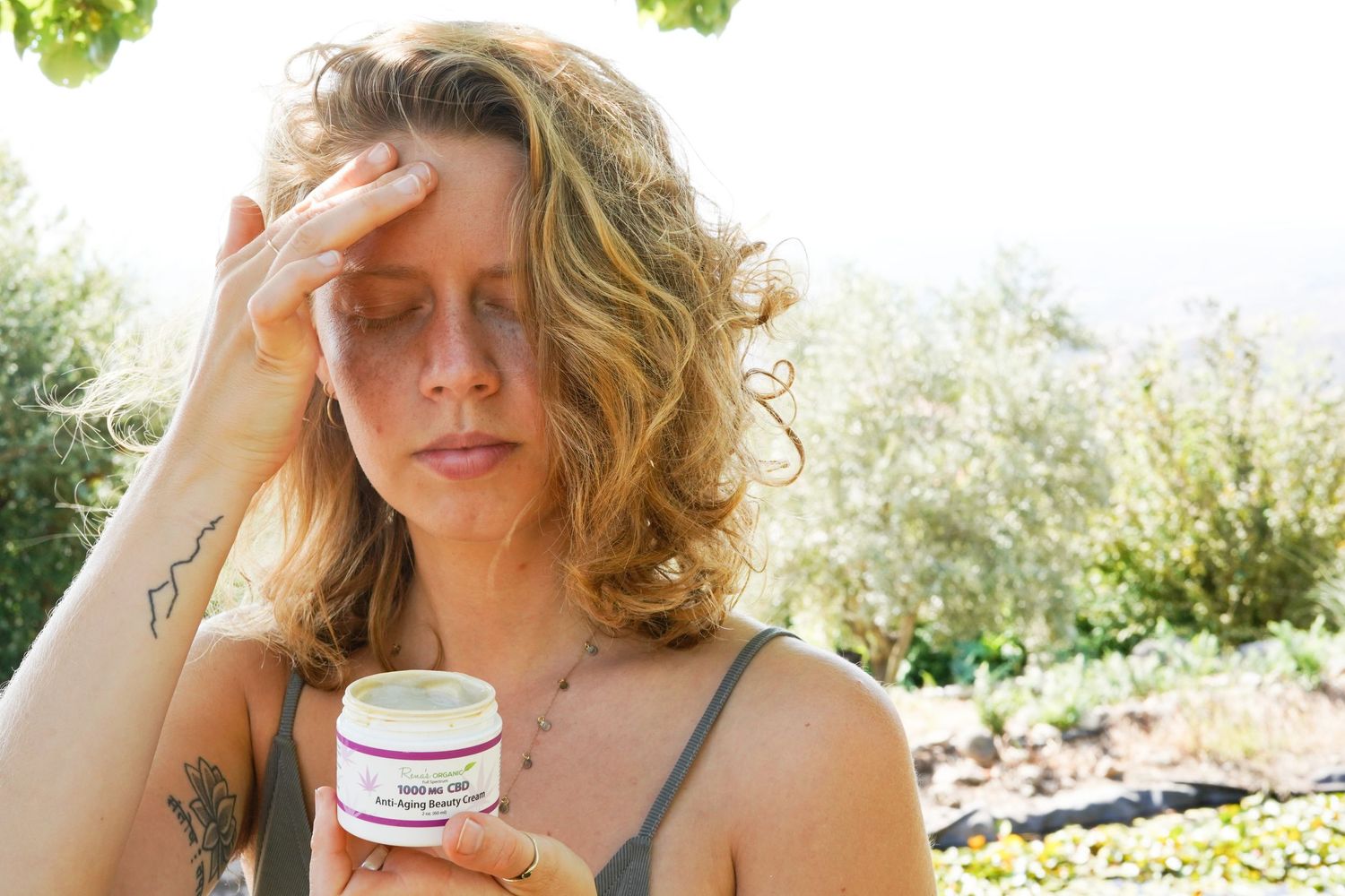 Woman holding a skincare cream jar outdoors, touching her forehead, with lush green garden background.