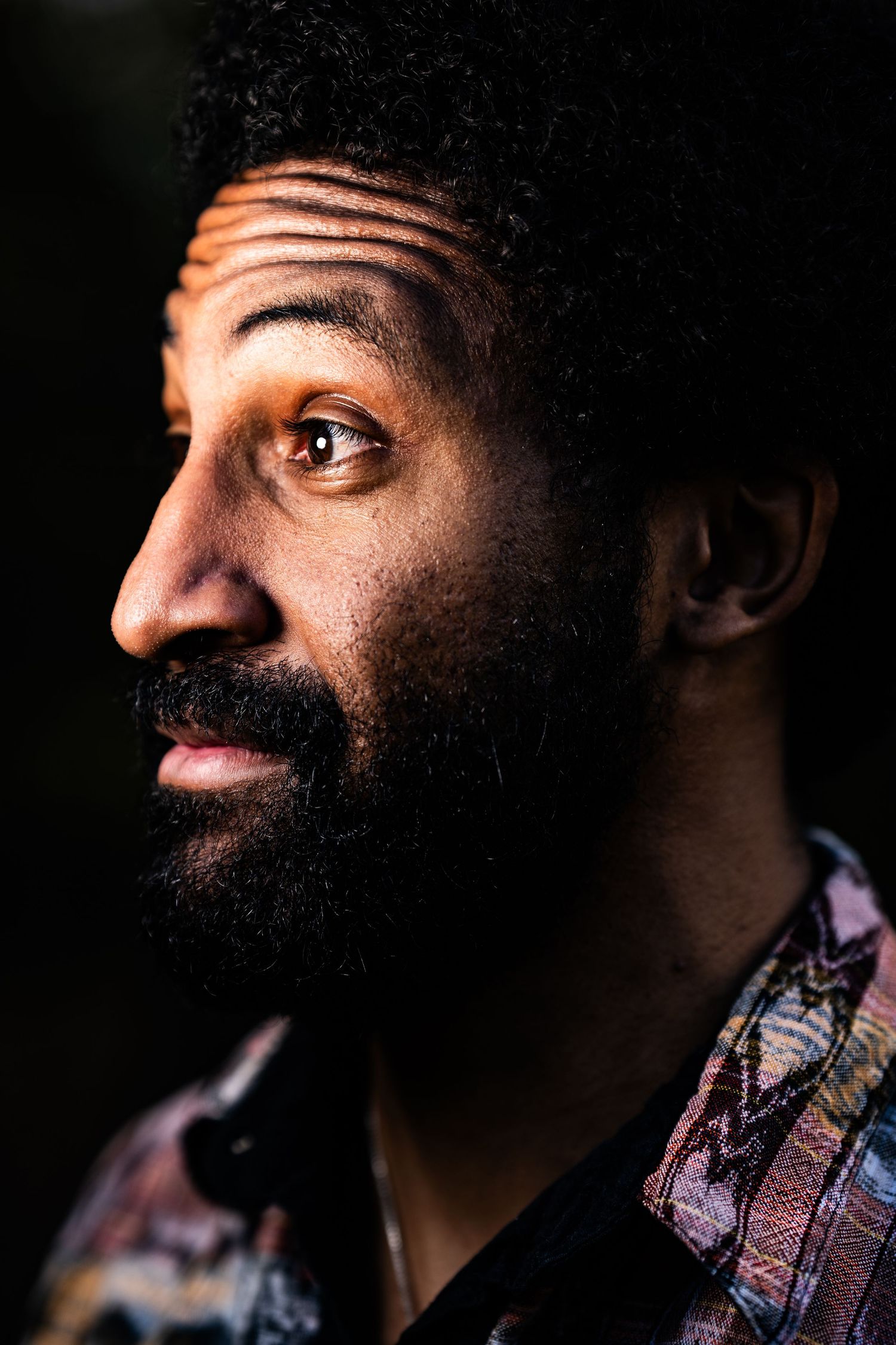 Close-up portrait of a bearded Black man in a colorful patterned shirt, gazing upward against a dark background.