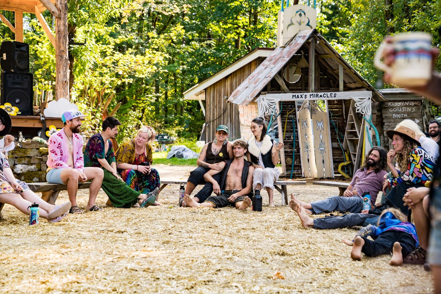Group of friends relaxing outdoors at a rustic festival campsite surrounded by trees and a wooden shack.