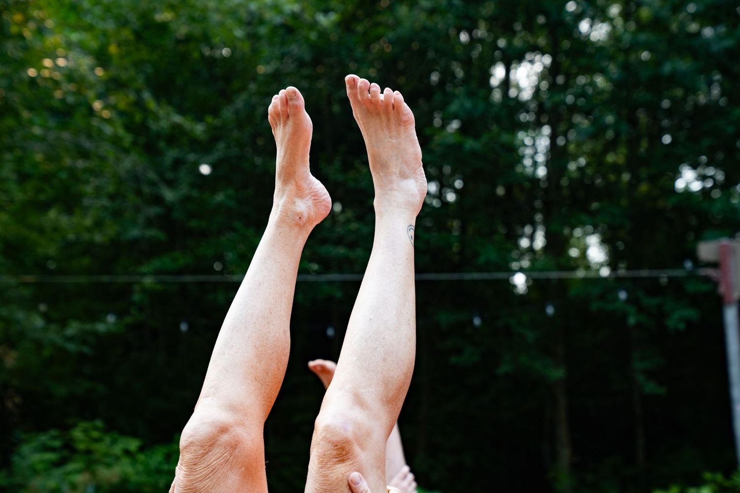 Two pairs of bare legs raised up in the air outdoors with green trees in the background.