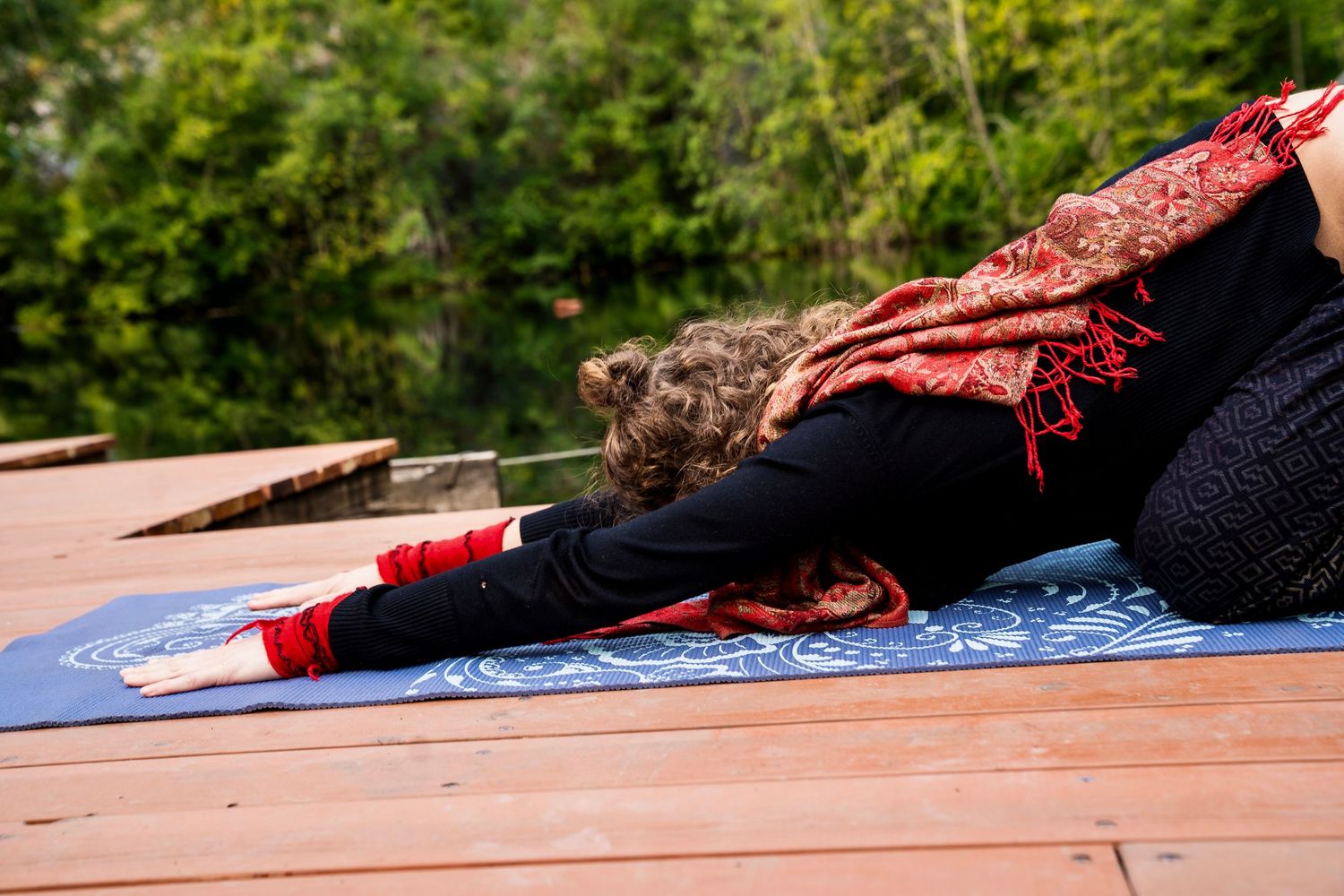Woman practicing yoga in child's pose on a blue mat outdoors on a wooden deck surrounded by green trees.