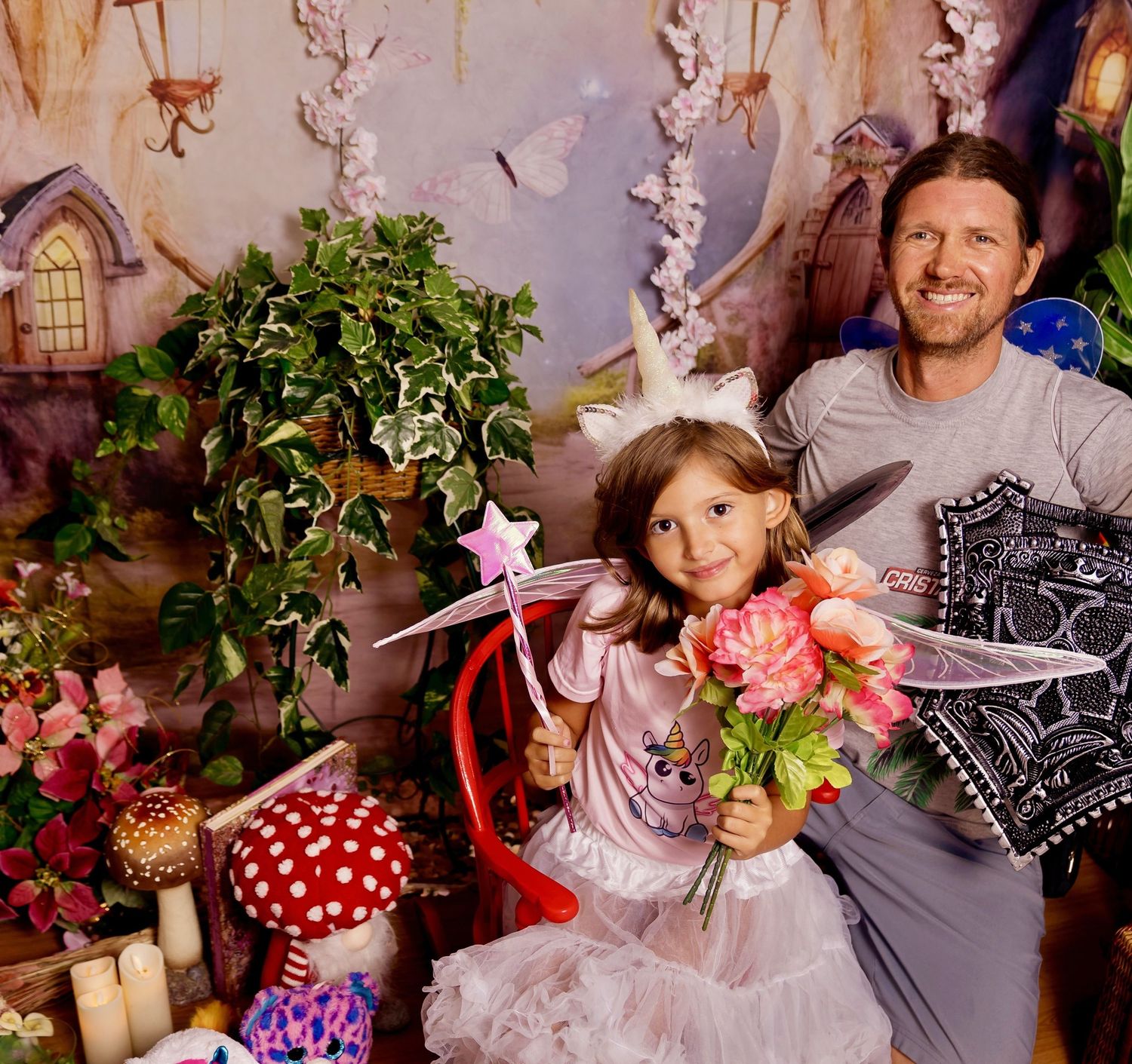 A man and young girl in fairy costume with wand and flowers pose before an enchanted forest backdrop.