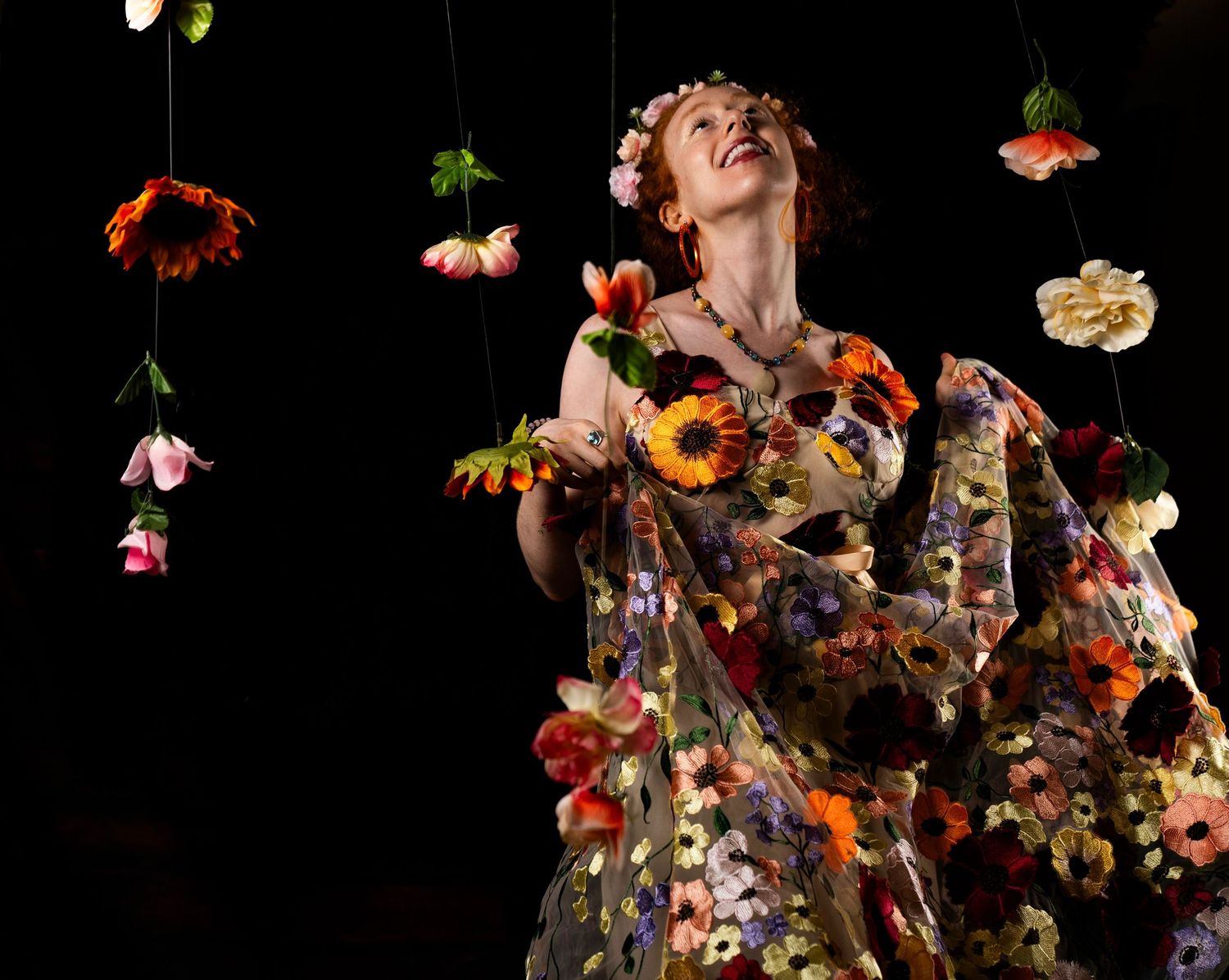 Woman in floral dress gazes upward surrounded by hanging flowers against dark background.
