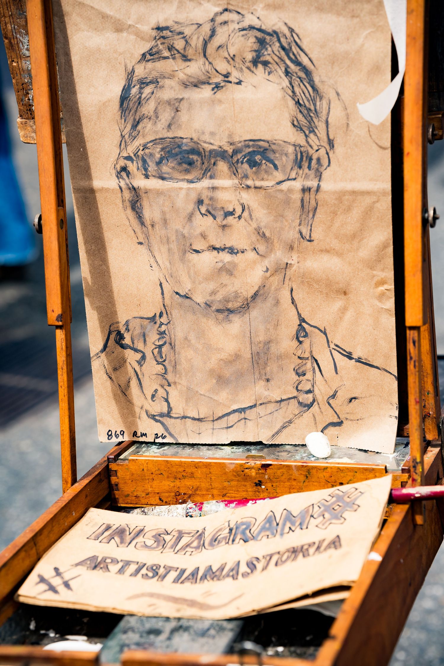 Street artist sketch portrait on paper displayed on wooden easel with Instagram handle written below.