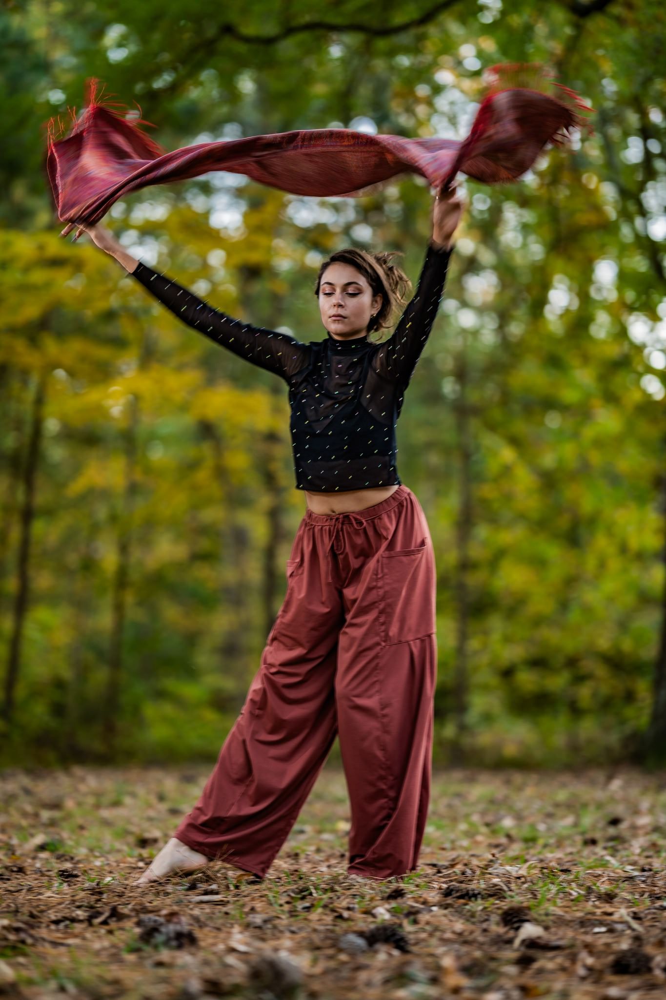 Young woman in red wide-leg pants and black top dancing barefoot in autumn forest holding flowing fabric.