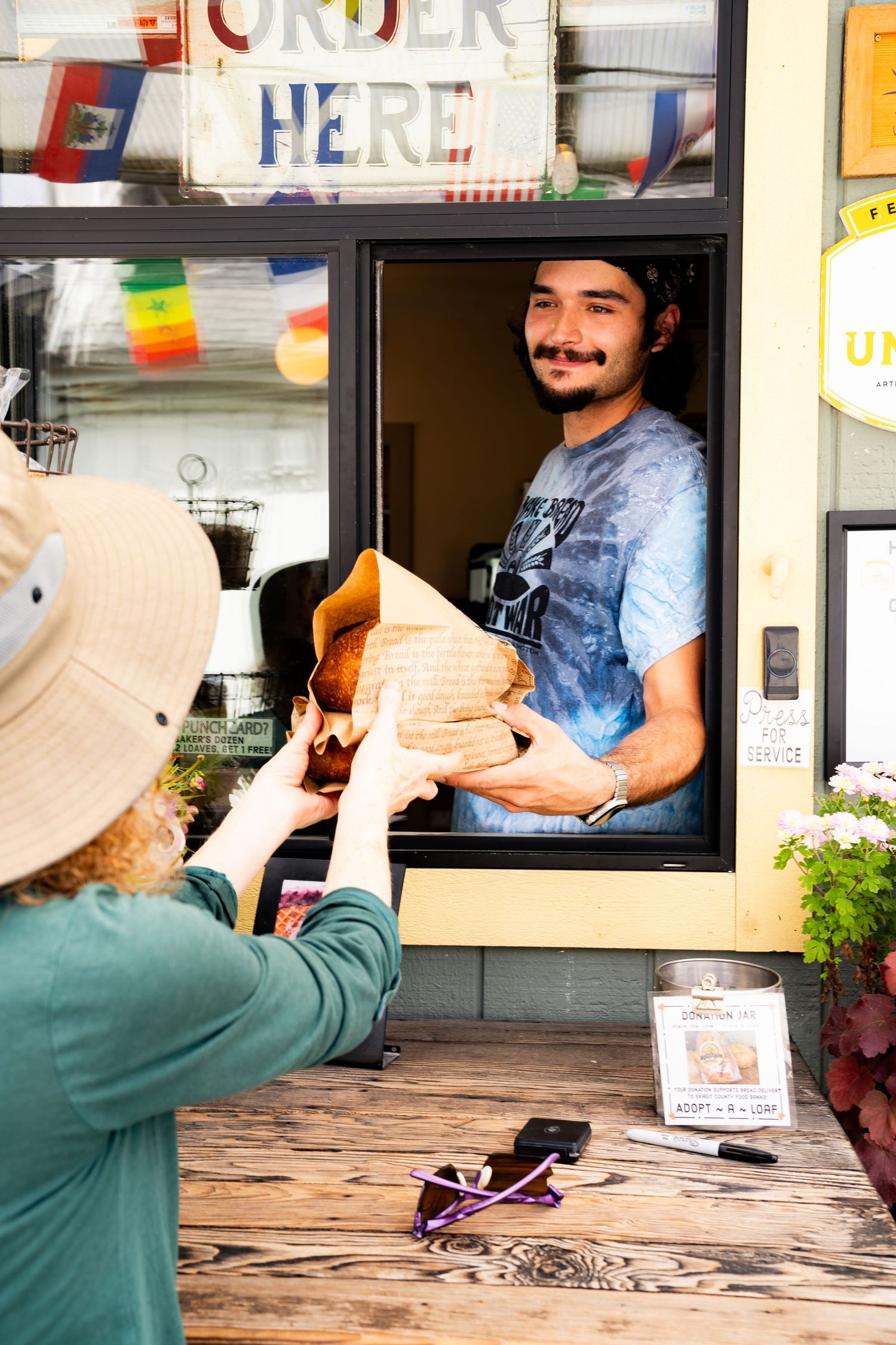 Young man with mustache handing brown paper bag to customer at food service window with Order Here sign.