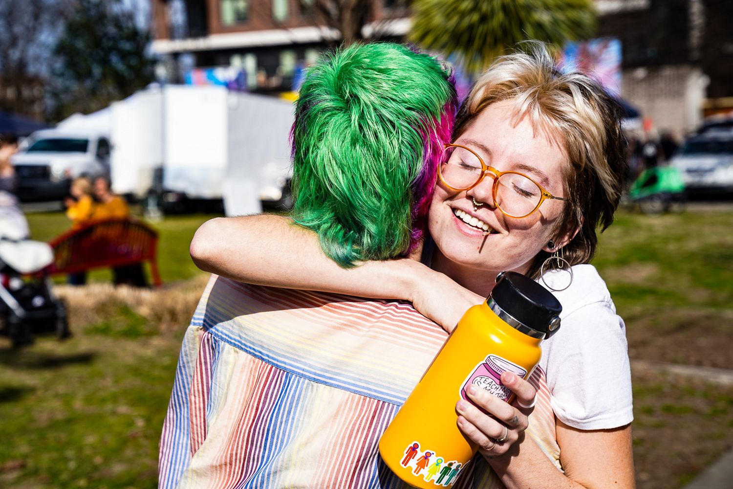 Two friends with short hair share a warm hug in downtown Boulder, one smiling with a toothpick in their mouth.