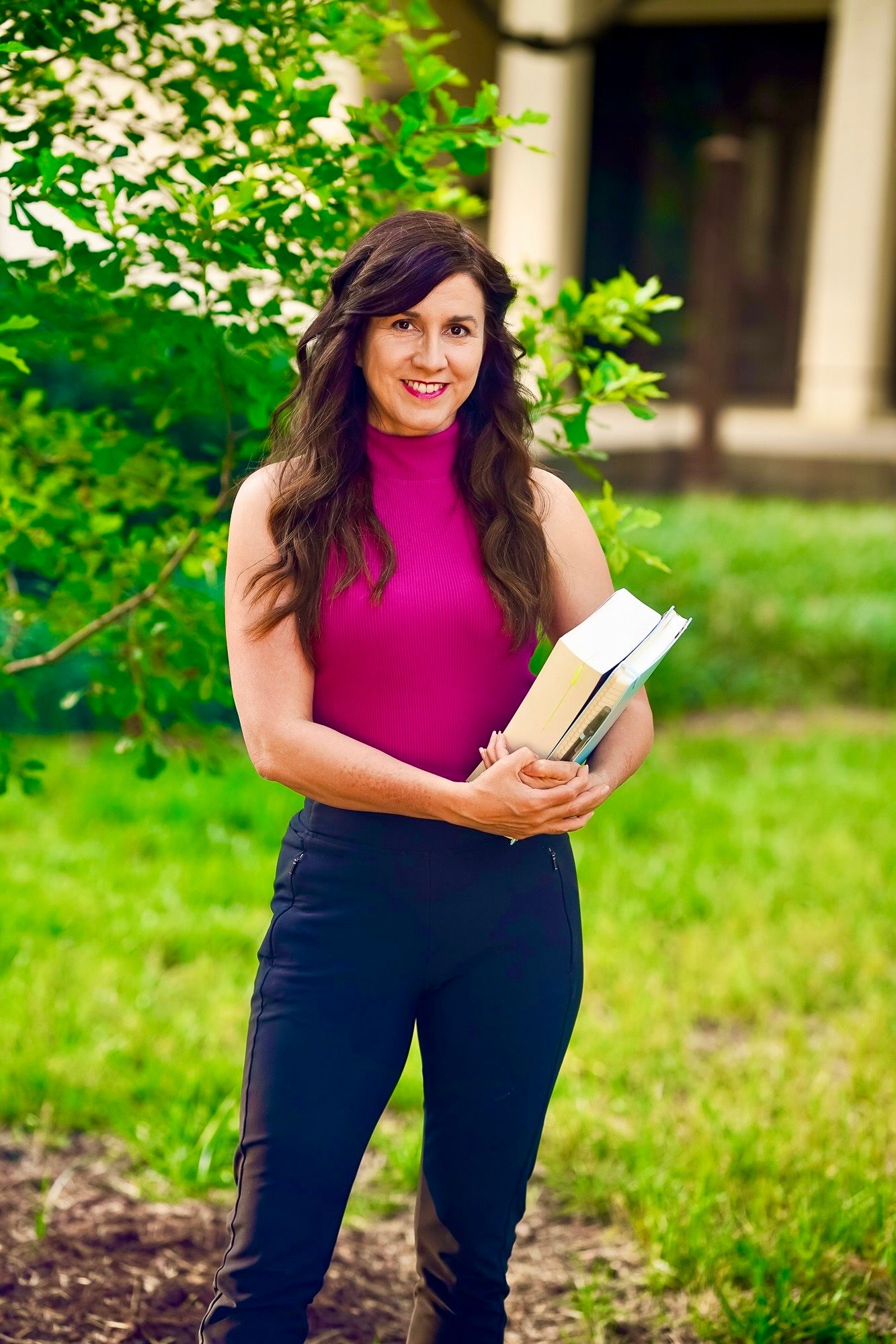 A woman in a bright pink sleeveless top and dark pants reads a book while standing in a lush green garden setting.