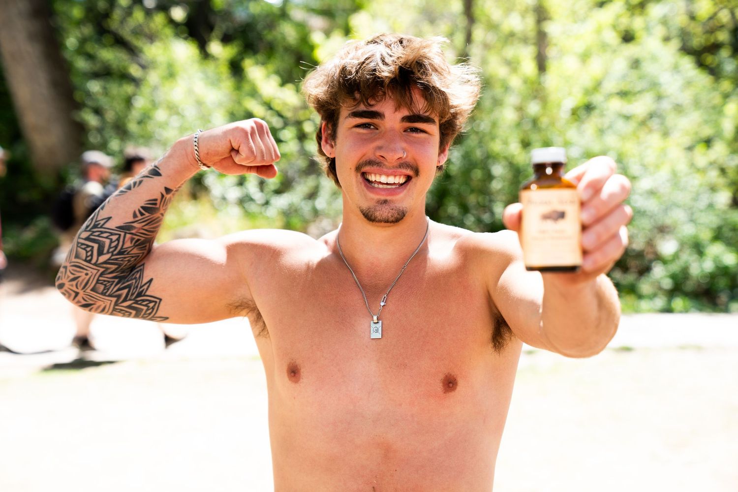 Smiling shirtless young man with tattoo flexing arm while holding a small amber bottle outdoors.