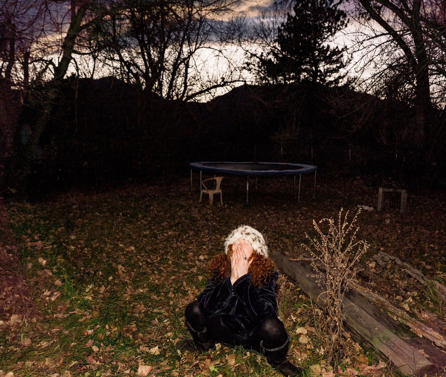 A trampoline sits in a backyard at dusk with fallen autumn leaves scattered on the ground.