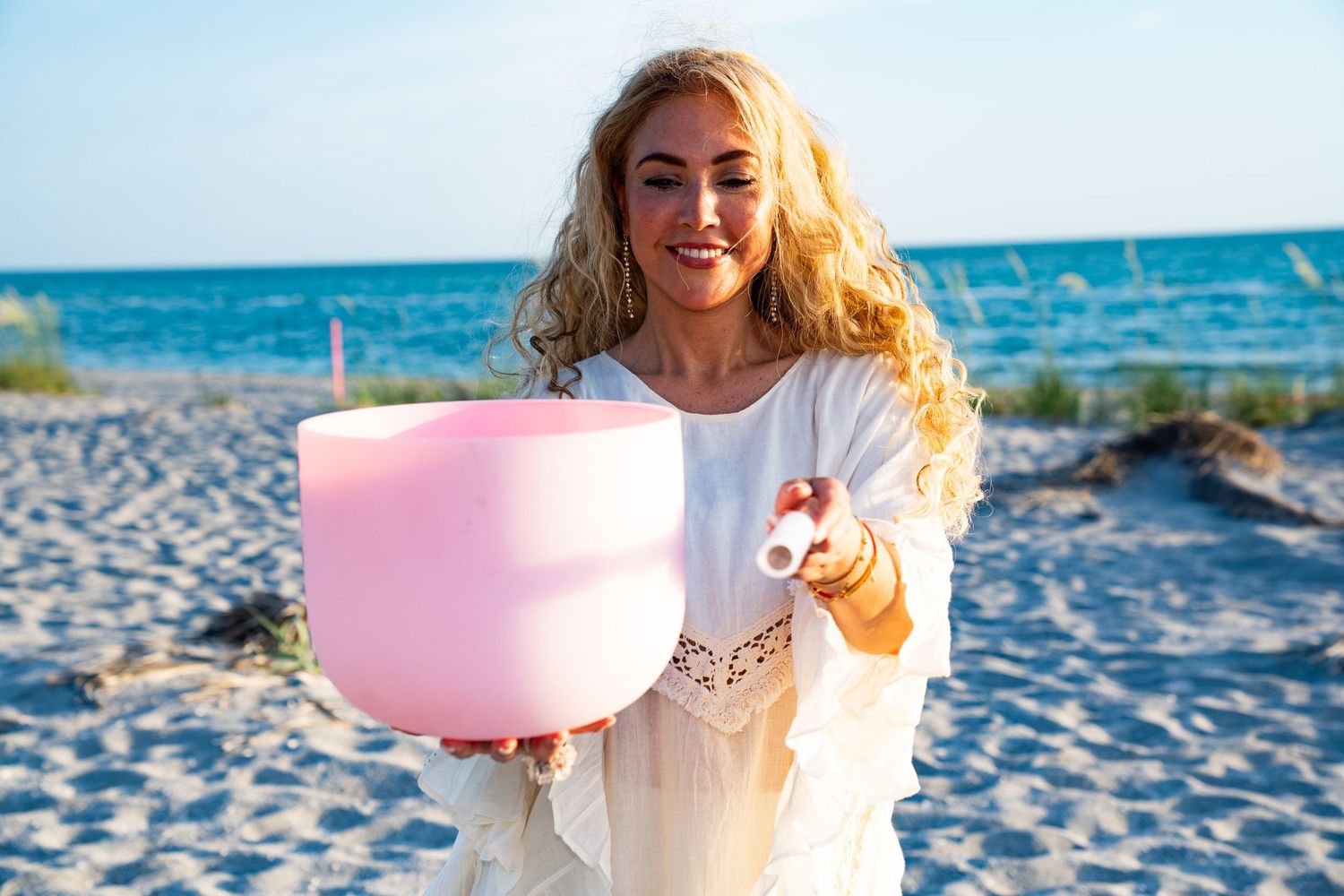 A photo of a woman who is blonde and is on the beach holding a pink singing bowl.