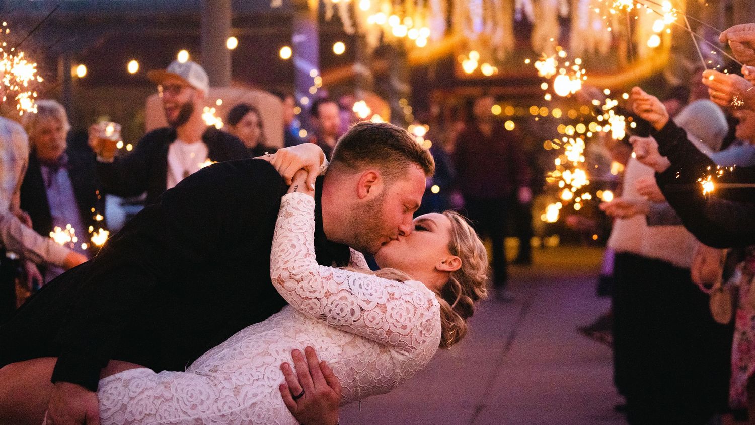 Couple sharing a romantic dip kiss at wedding reception surrounded by guests holding sparklers at night.