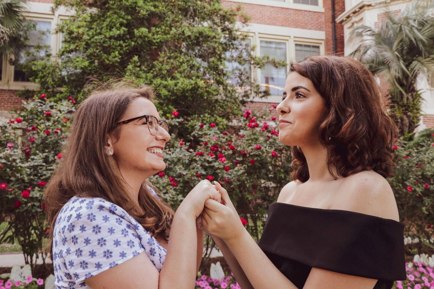 Two young women holding hands and smiling at each other outdoors with colorful flowers and greenery in the background.