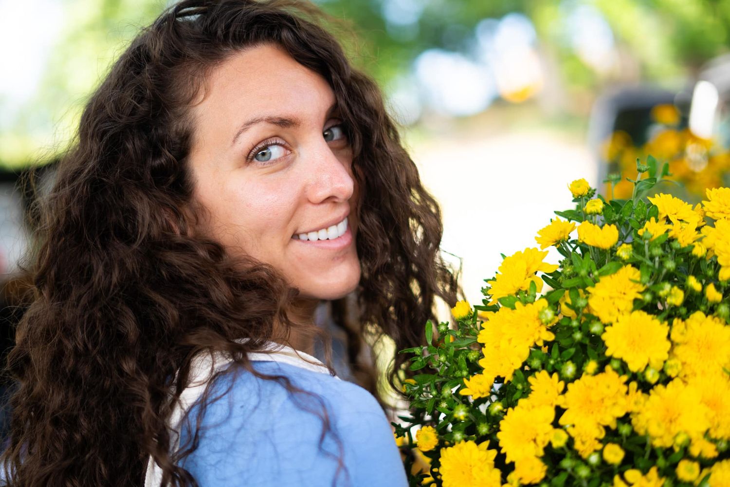 Smiling woman with curly brown hair holding vibrant yellow chrysanthemum flowers outdoors.