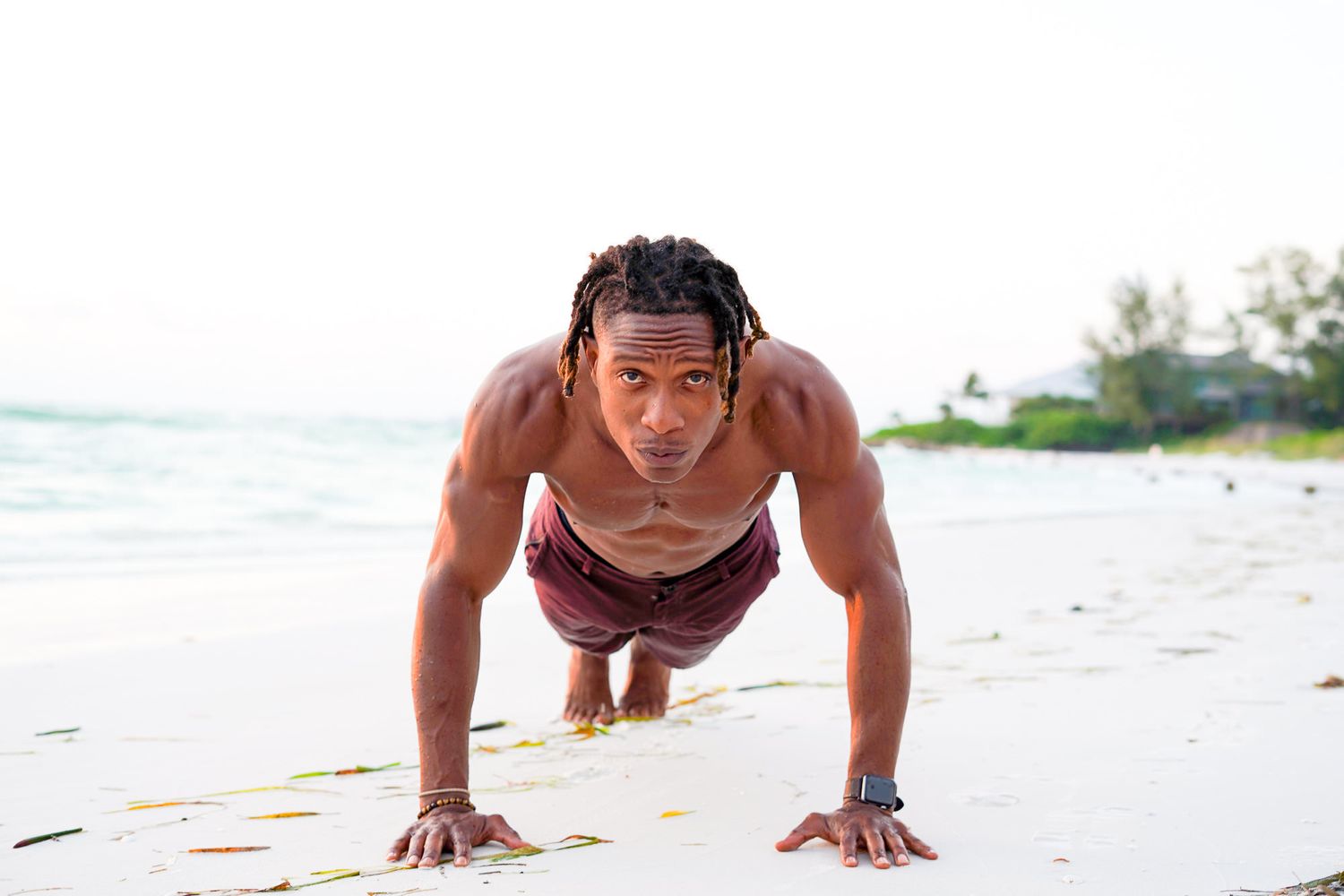 Muscular man doing push-ups on sandy beach near ocean during outdoor workout session.