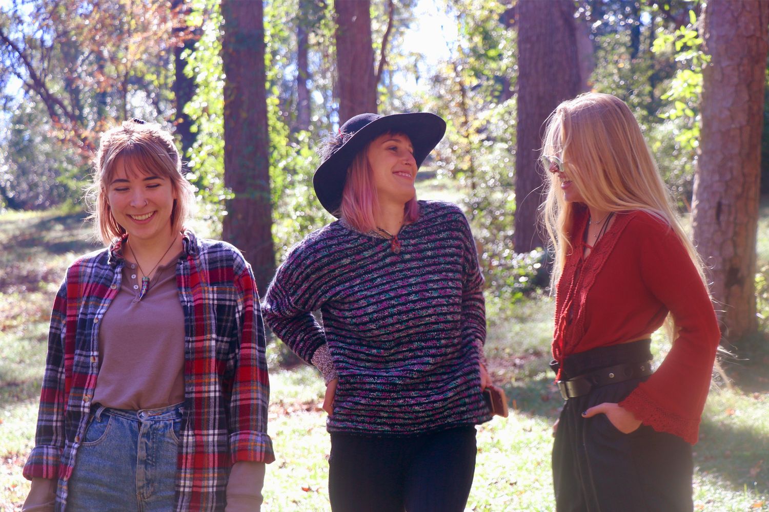Three women smiling and chatting outdoors in a sunlit forest setting, wearing casual autumn outfits.