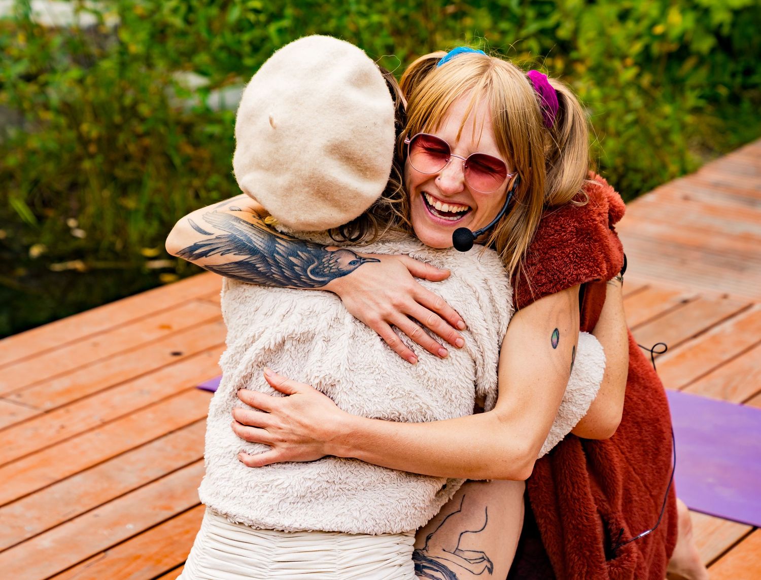 Two tattooed women hugging warmly outdoors on a wooden deck, smiling joyfully in casual boho outfits.