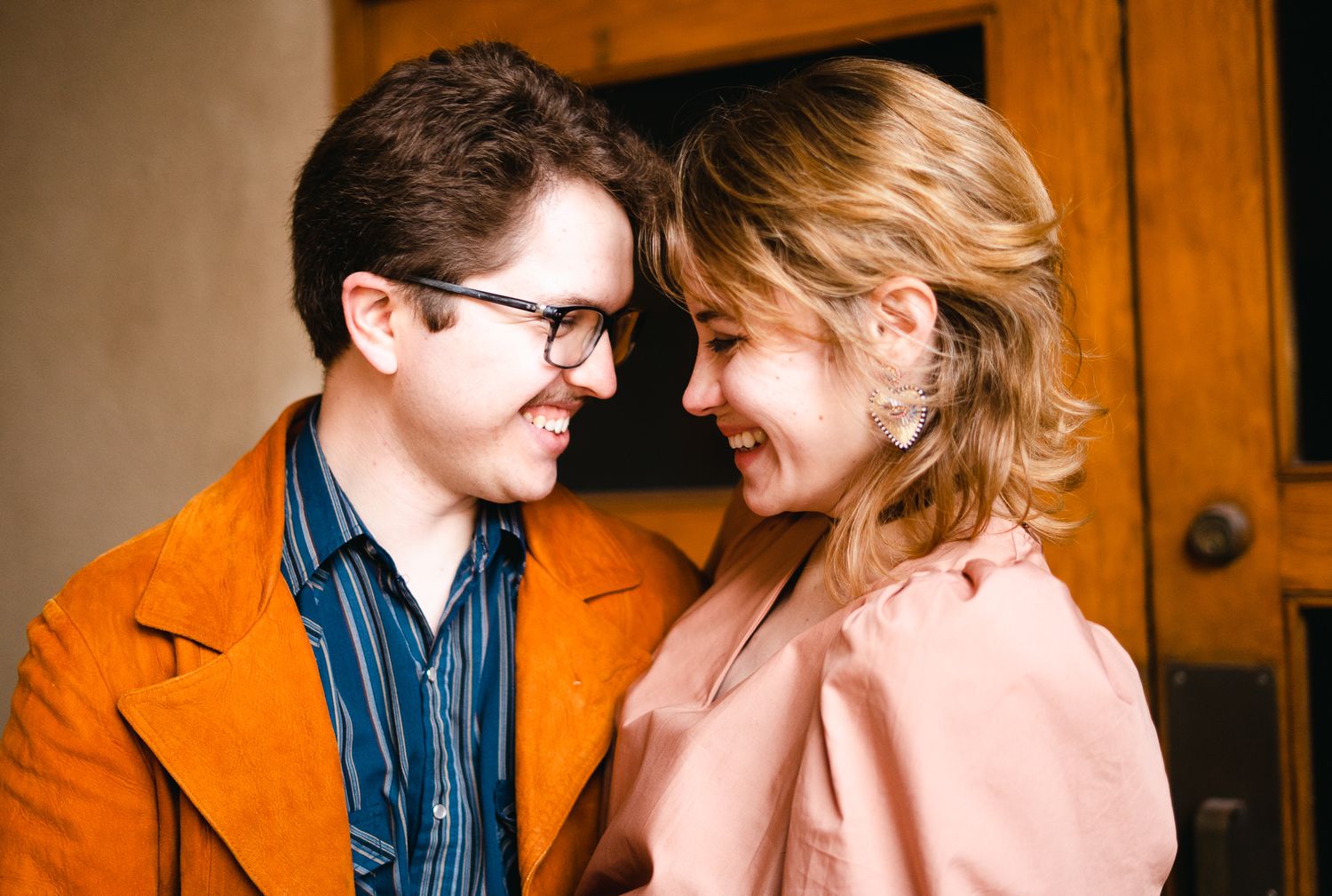 A smiling couple in colorful jackets share a tender moment, gazing at each other near a wooden door.