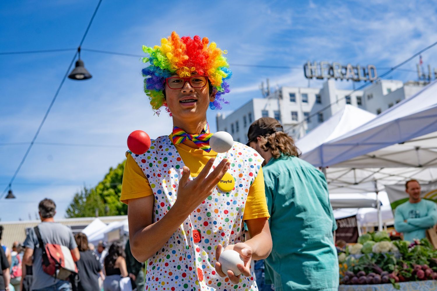 Colorful clown in rainbow wig juggling at an outdoor farmers market on a sunny day.