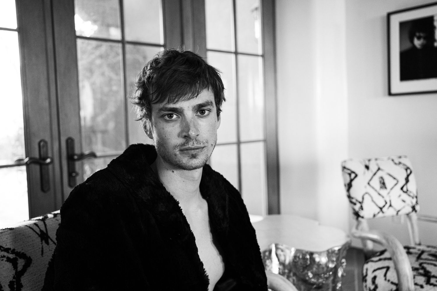 Brooding young man in black robe seated indoors in black and white portrait photo.
