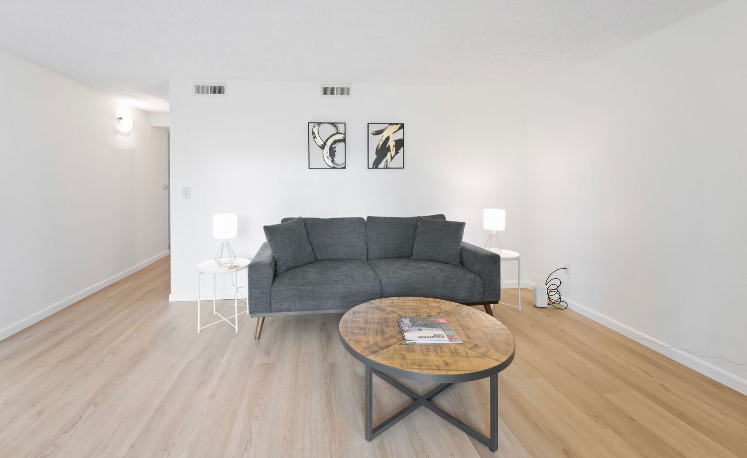 Modern living room with gray sofa, rustic wood coffee table, hardwood floors, and minimalist white walls with framed art.