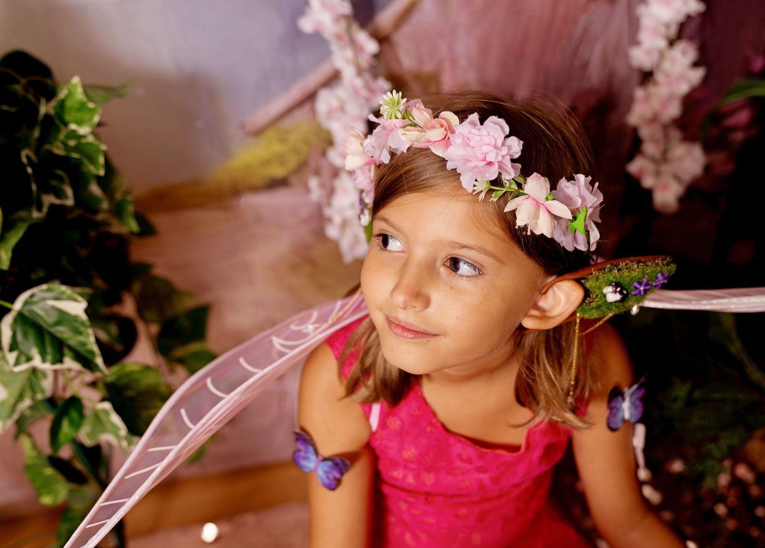 Young girl dressed as a fairy with pink flower crown, butterfly wings, and pink dress in an enchanted garden setting.