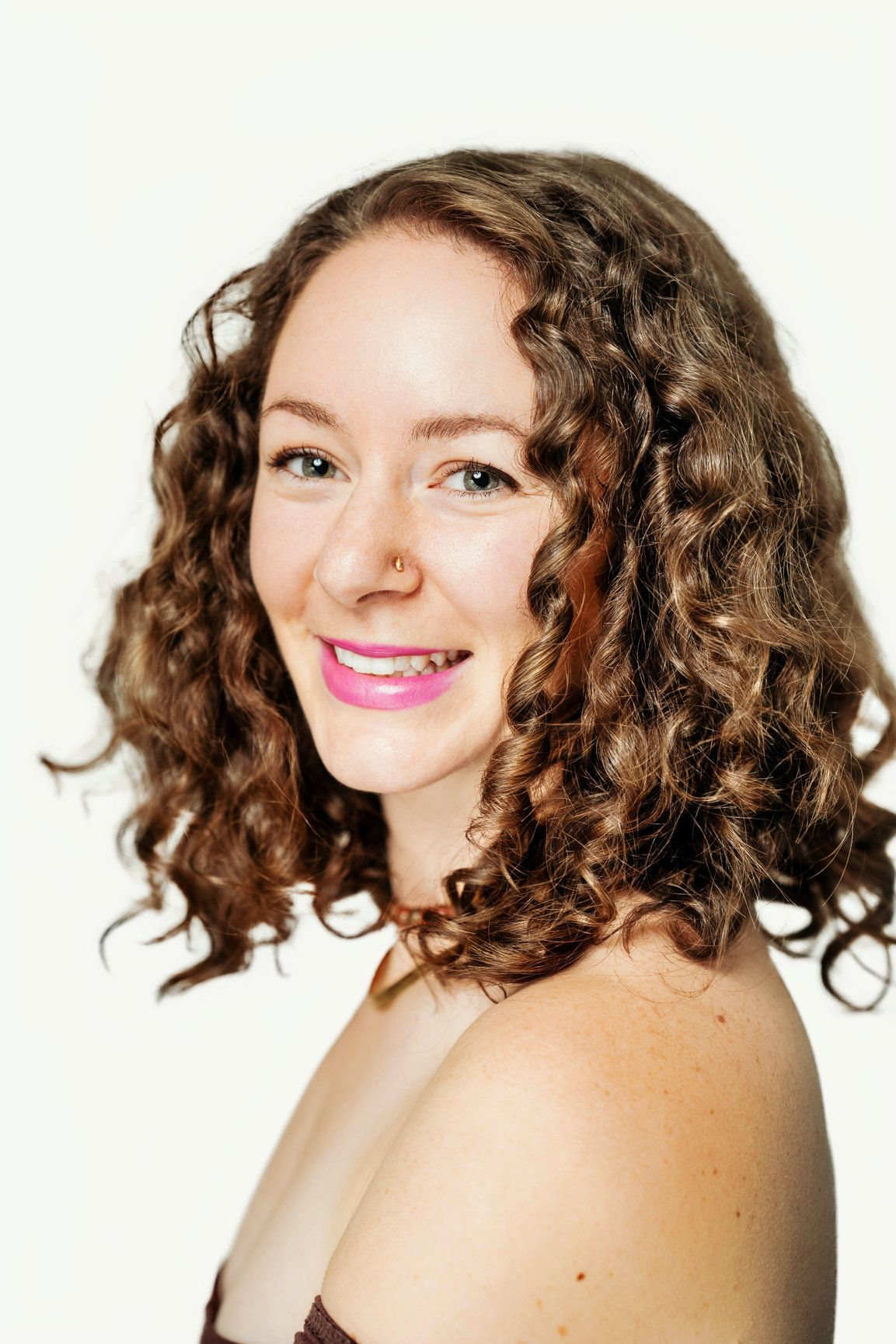 A woman is smiling at the camera, she has brown curly hair and is wearing a brown top. She is in a photography studio.