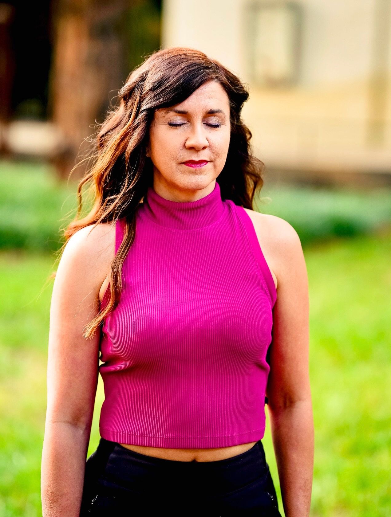 A photo of a woman with her eyes closed wearing a hot pink shirt and greenery behind her.