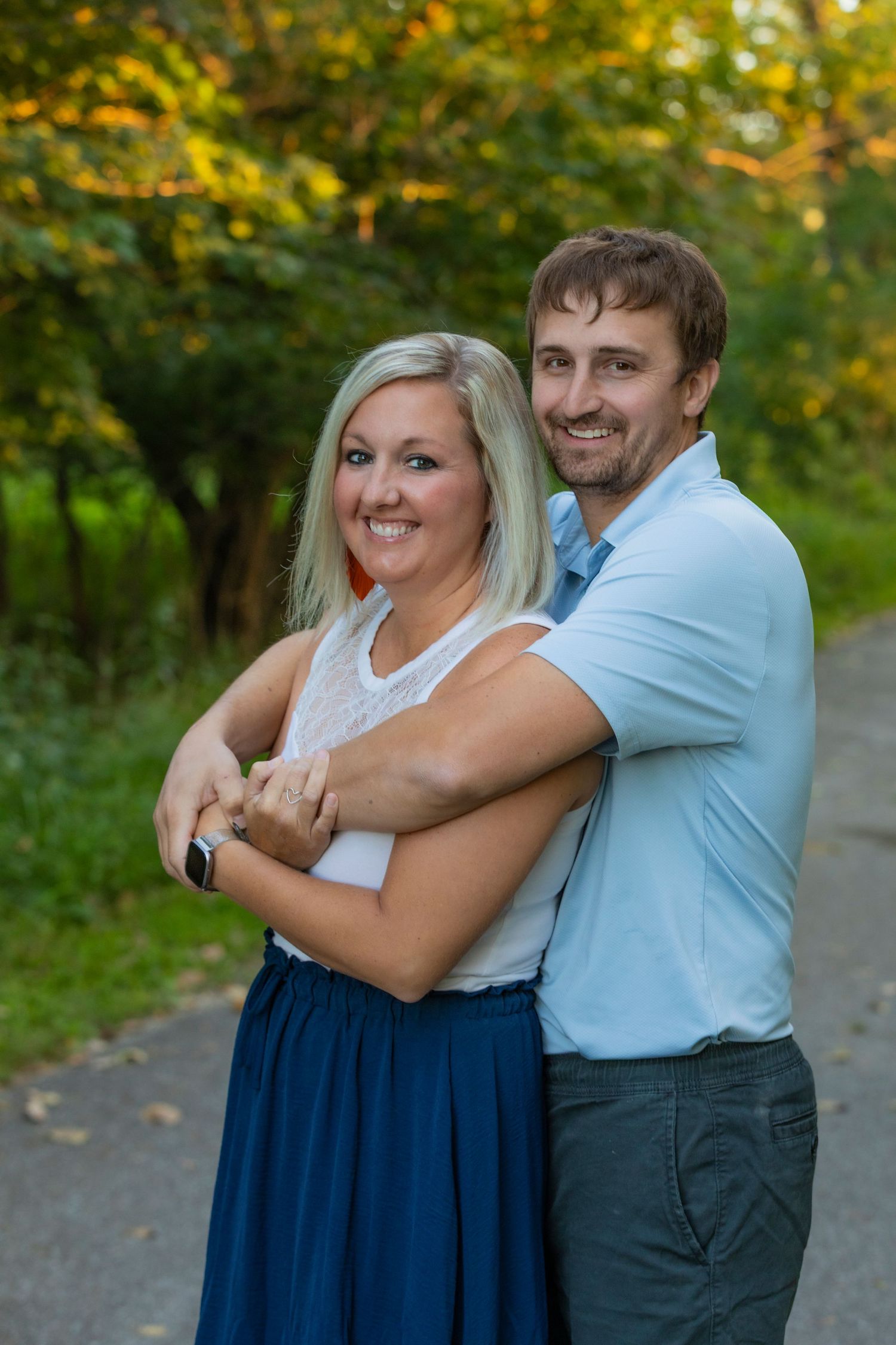 A couple embraces and laughs together on a path surrounded by trees with golden autumn foliage in the background.