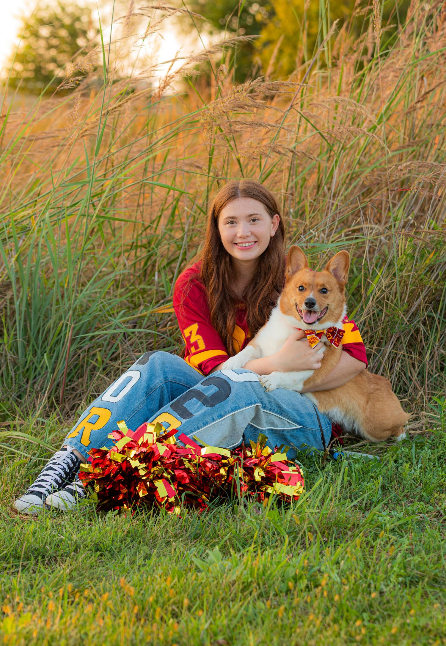 A corgi dog rests in autumn grass next to someone wearing red and yellow clothing with colorful fall flowers nearby.