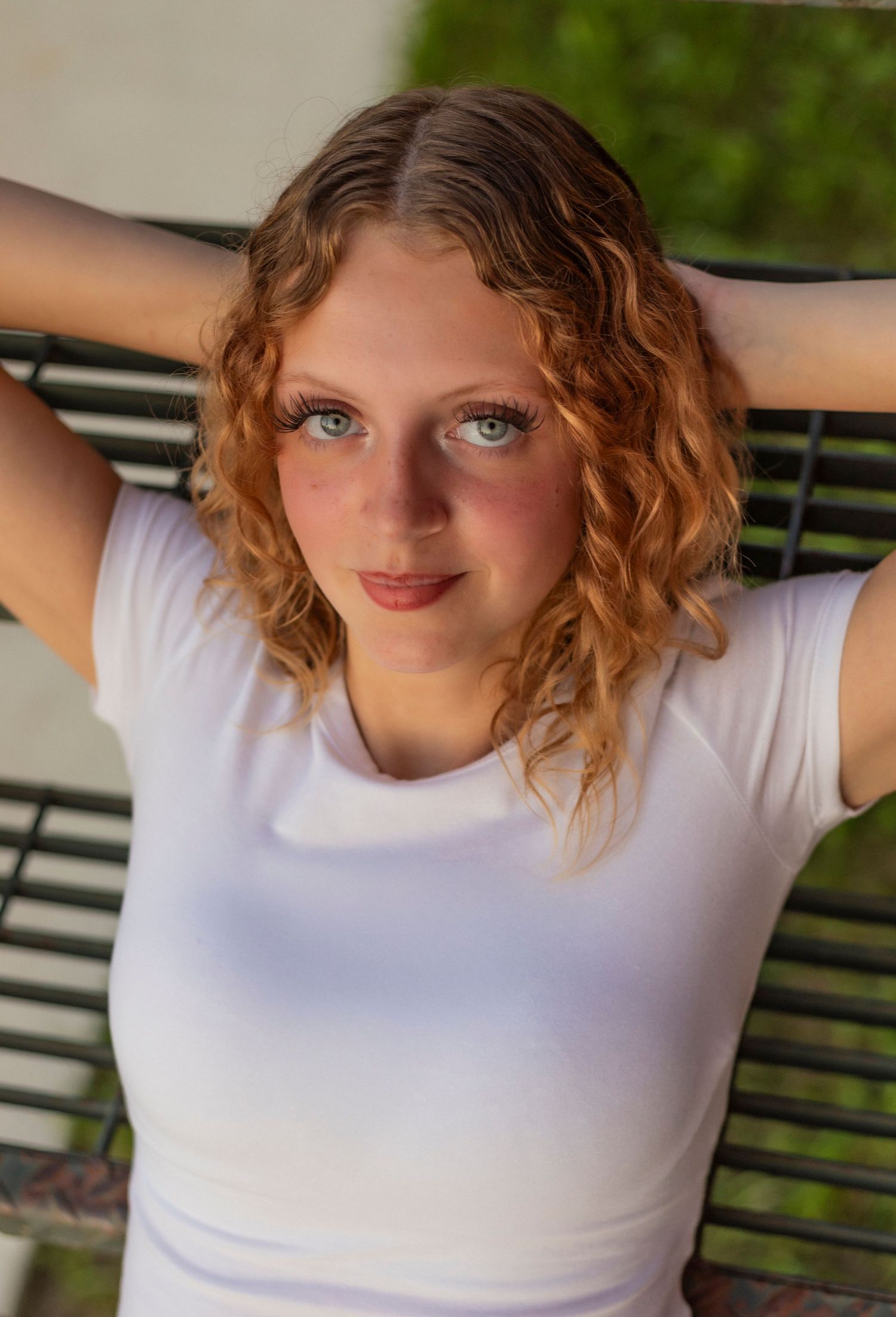 Young woman with curly red hair and green eyes poses on a bench outdoors in a white t-shirt.