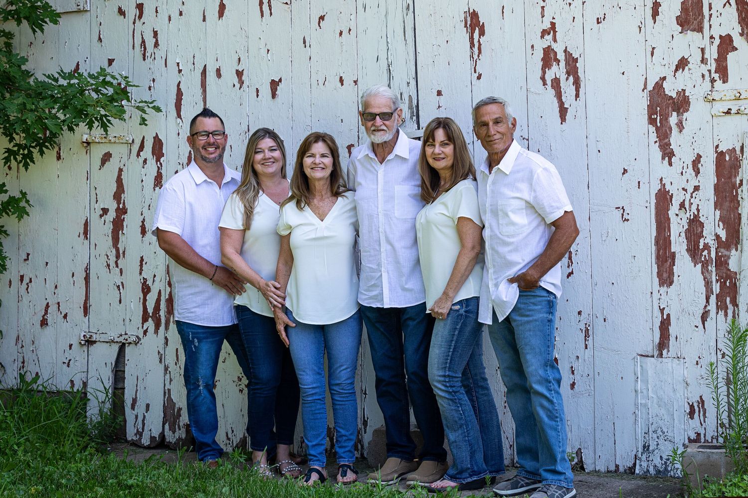 A group in coordinated white shirts and blue jeans poses casually against a rustic white barn wall in an outdoor setting.