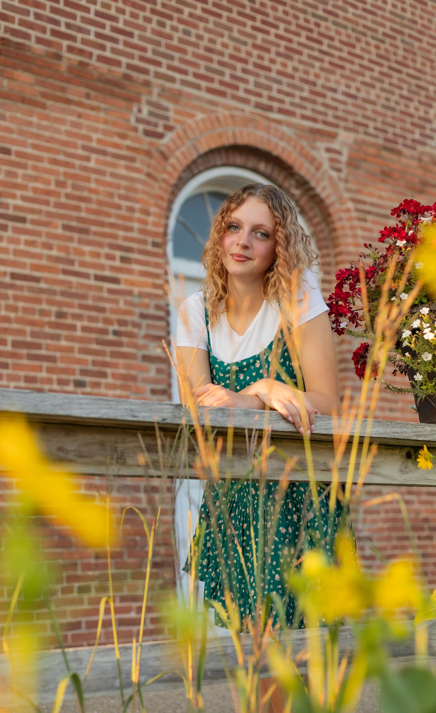 Young woman in green floral dress leans on wooden fence surrounded by colorful flowers against brick building.