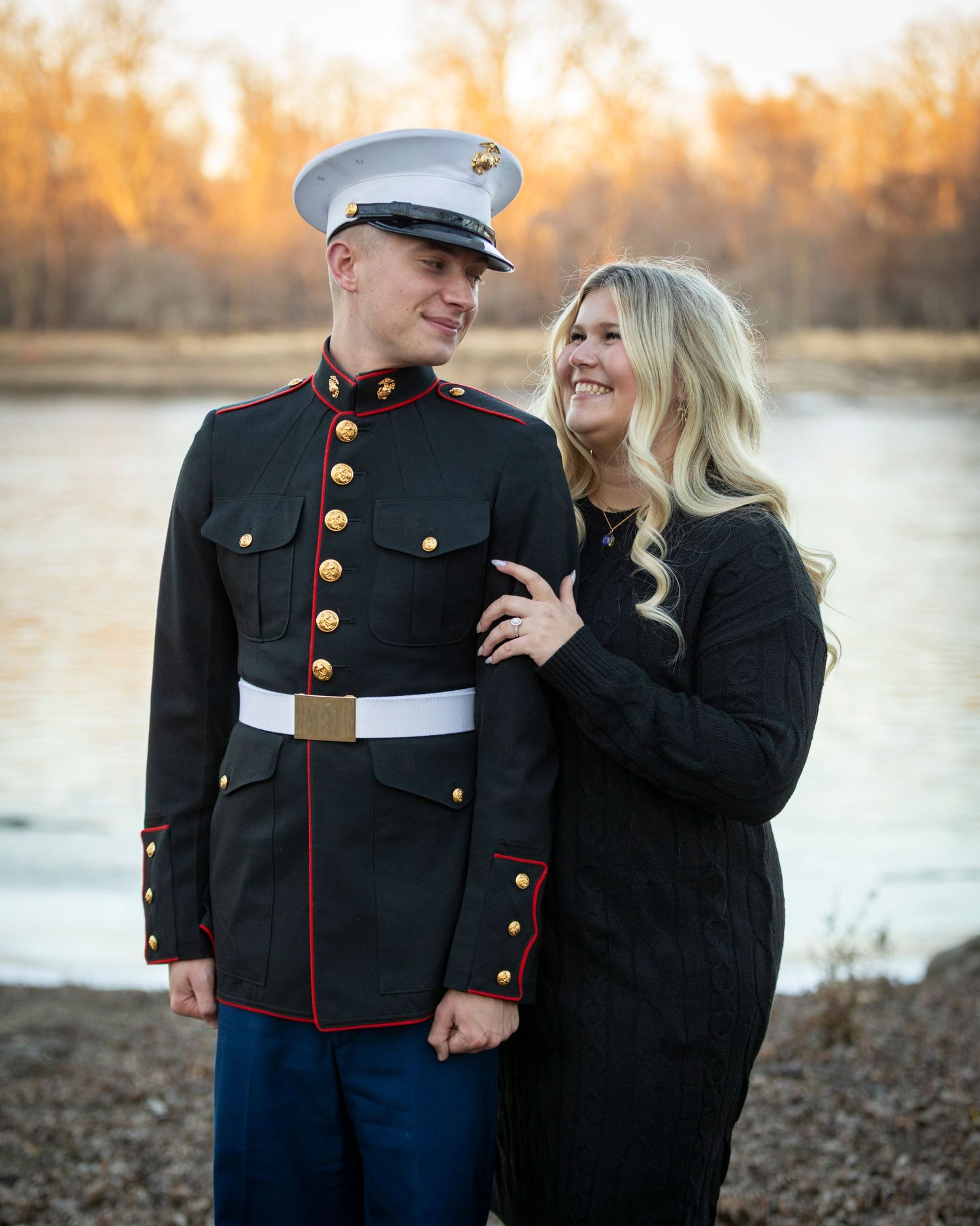 A couple poses together near a lake at sunset, with one person wearing a marine dress uniform and the other in black attire.