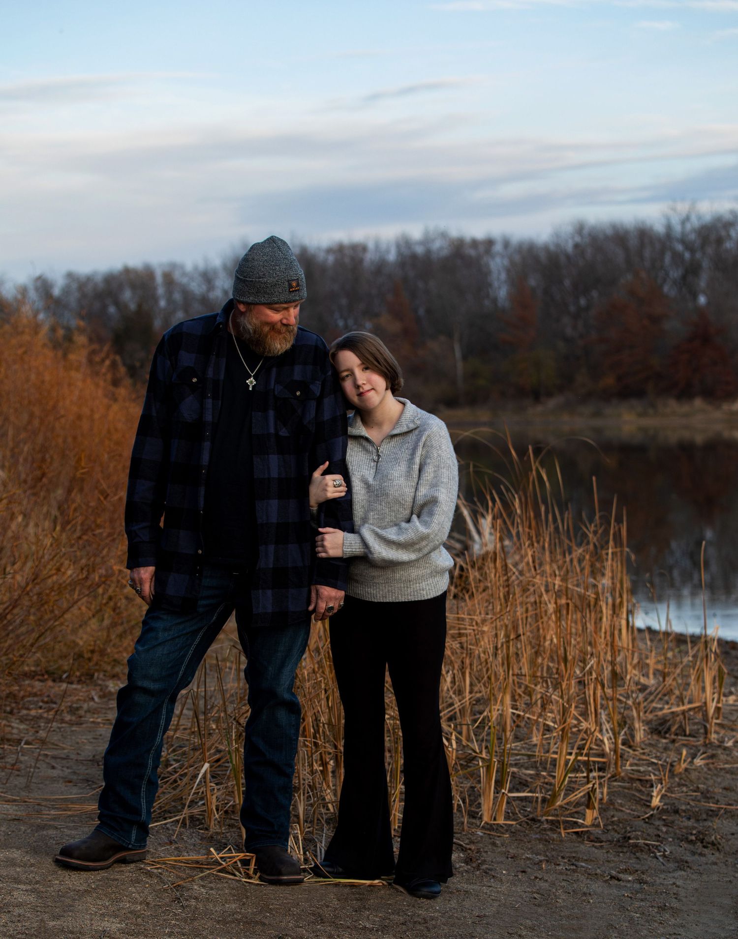 A couple stands together among tall reeds near a lake at sunset in a romantic outdoor setting.