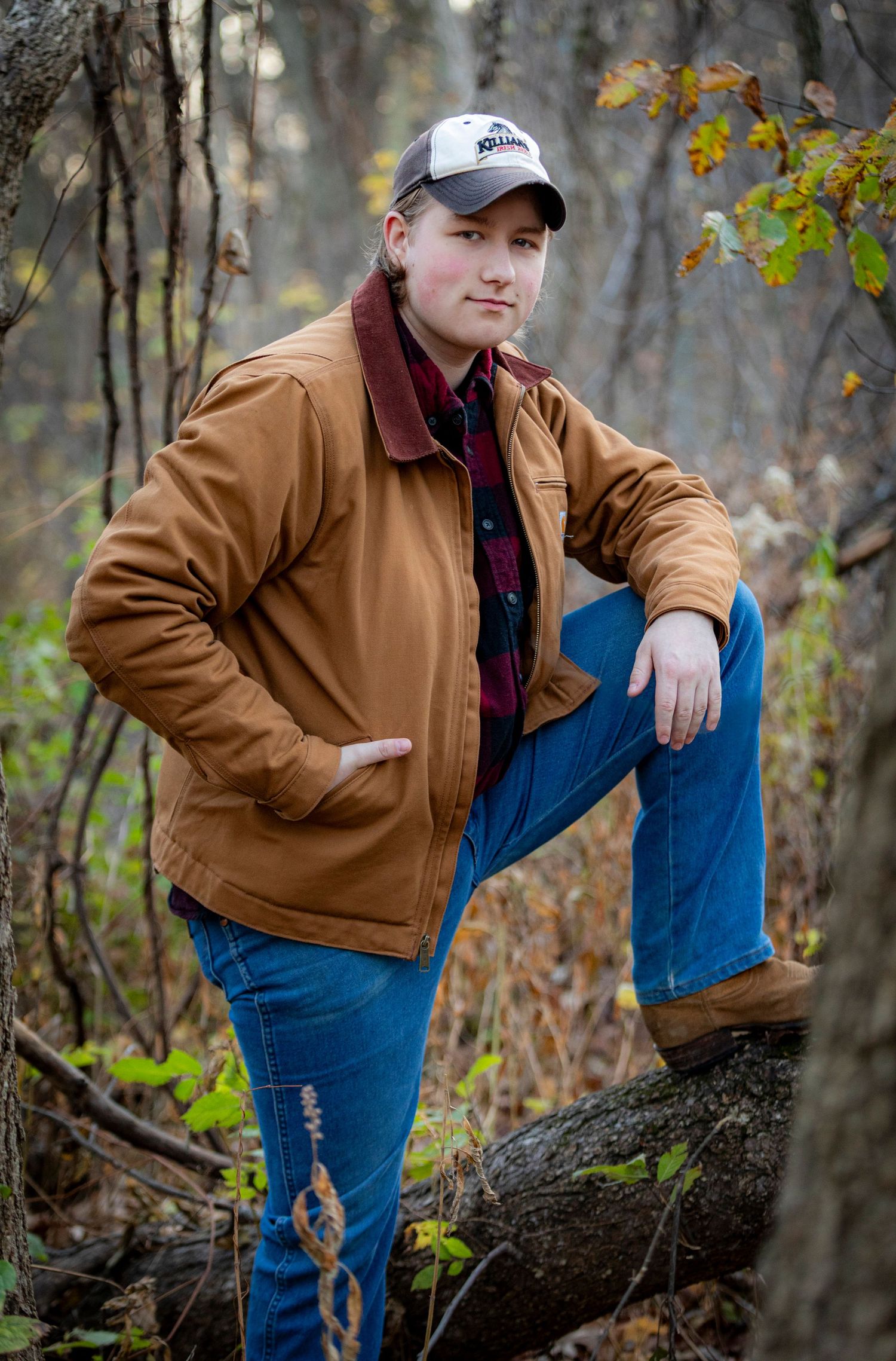 Person in brown jacket and jeans poses casually in autumn forest setting against fallen tree trunk.