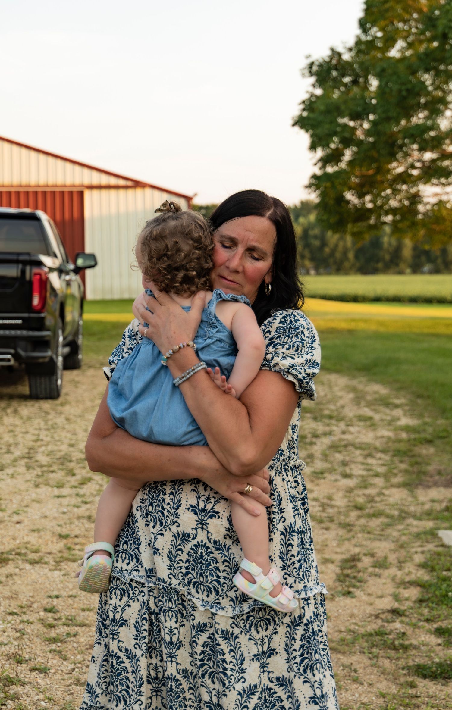 A tender embrace between grandparent and child in a blue dress and floral print dress against a rural outdoor setting.