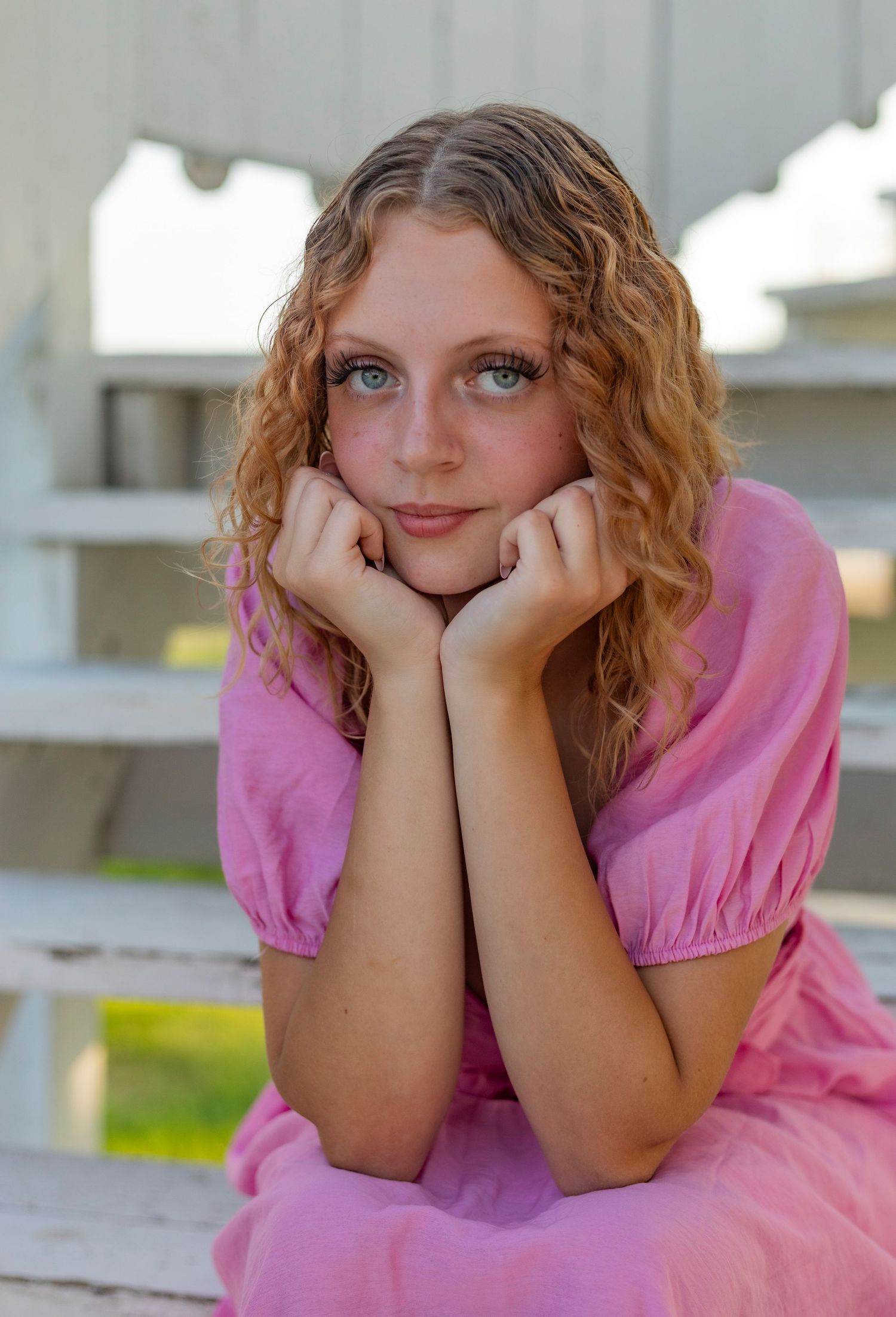 Young woman with curly blonde hair and blue eyes poses in a pink dress on white wooden bleachers outdoors.