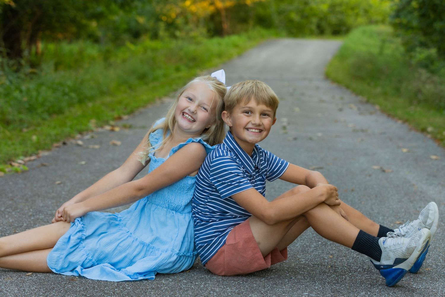 Brother and sister lying on a paved path surrounded by green foliage on a sunny day.