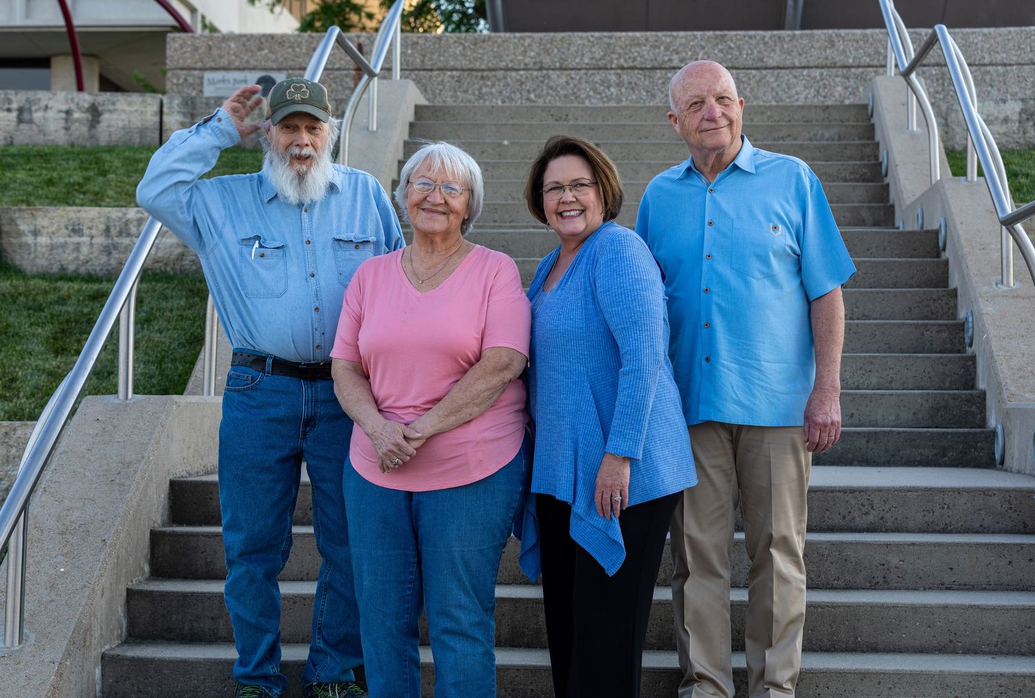 Four people stand together smiling on outdoor concrete steps with metal railings on a sunny day.