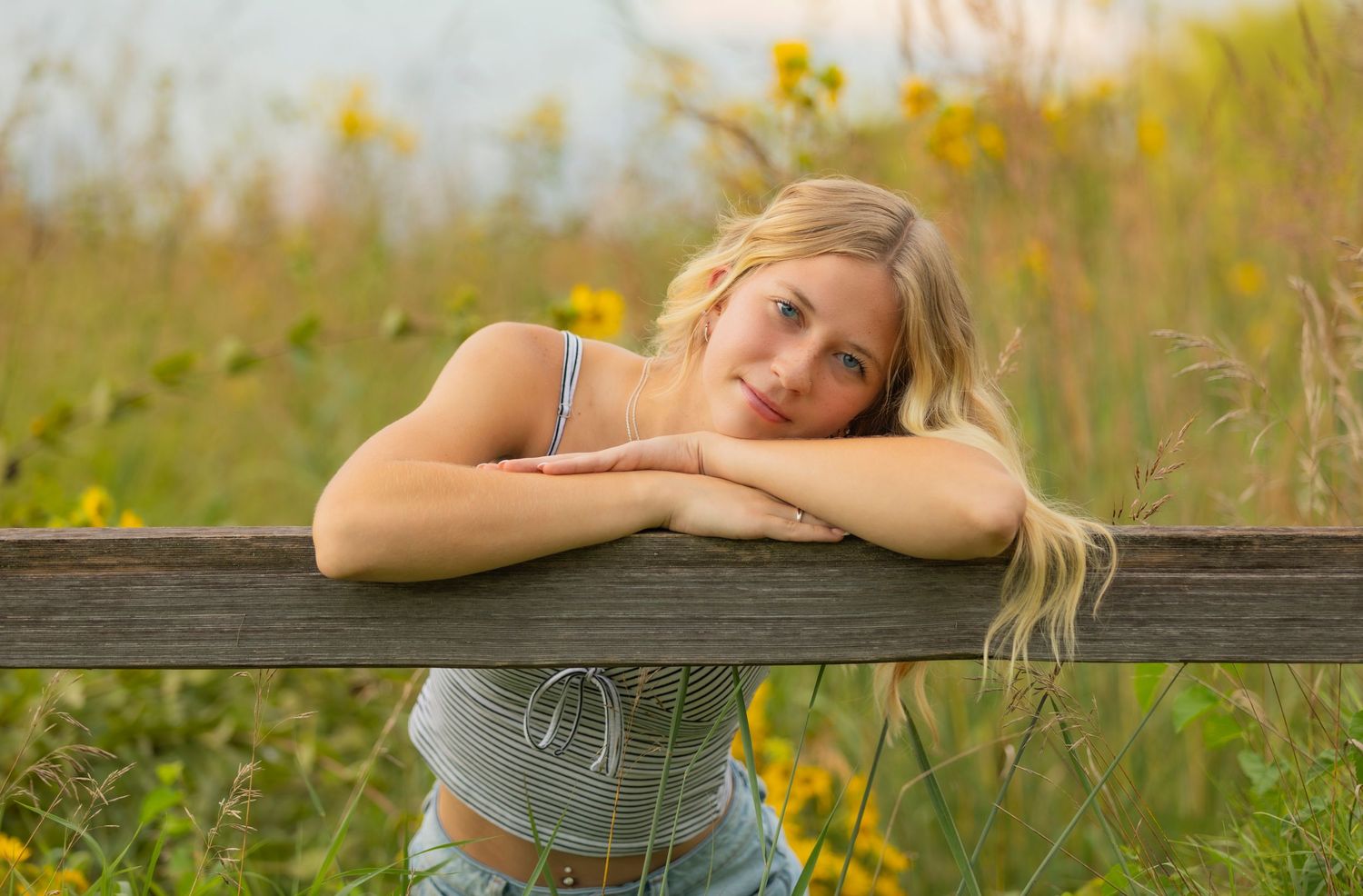 Woman in casual attire relaxes on wooden railing surrounded by wildflowers in a golden summer meadow.