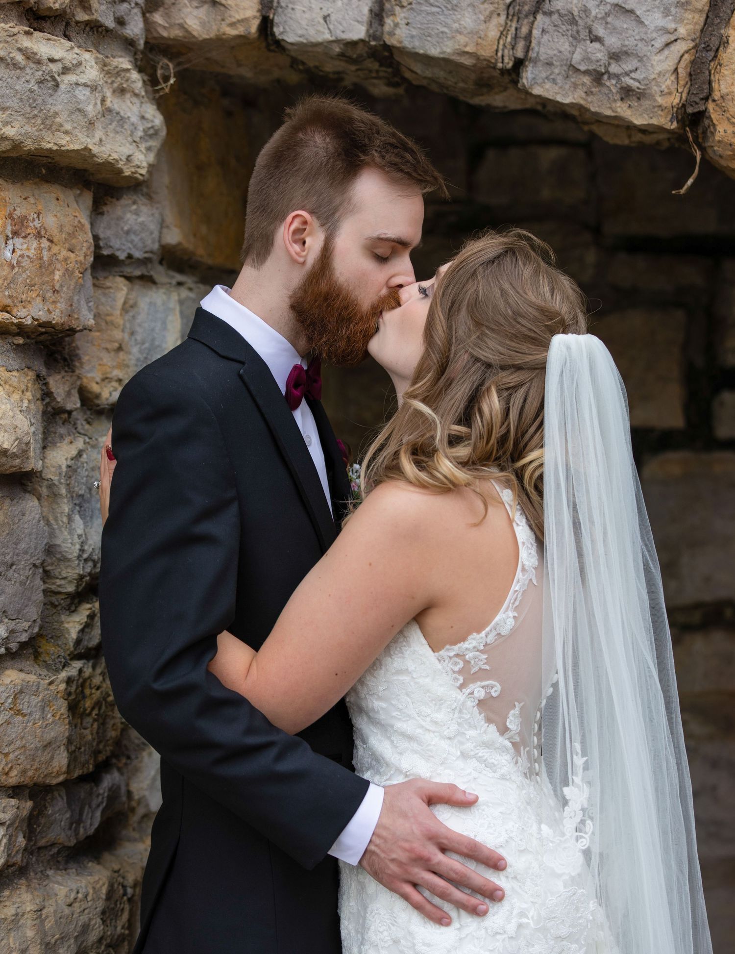 A romantic wedding couple embraces against a rustic stone wall backdrop while wearing formal attire.