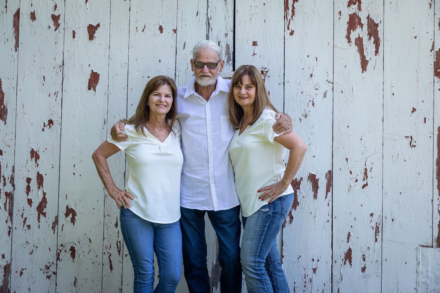 Three people in casual white tops and jeans pose together against a weathered white wooden wall background.