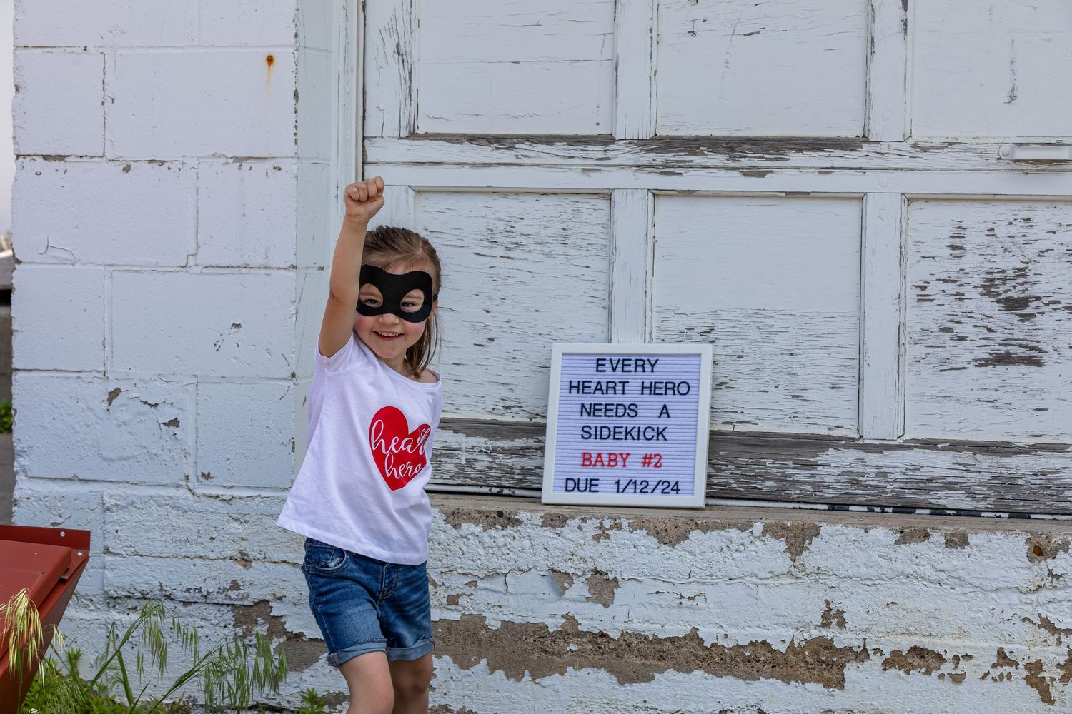 A child in a superhero mask and heart t-shirt stands by a white wall with a posted sign.