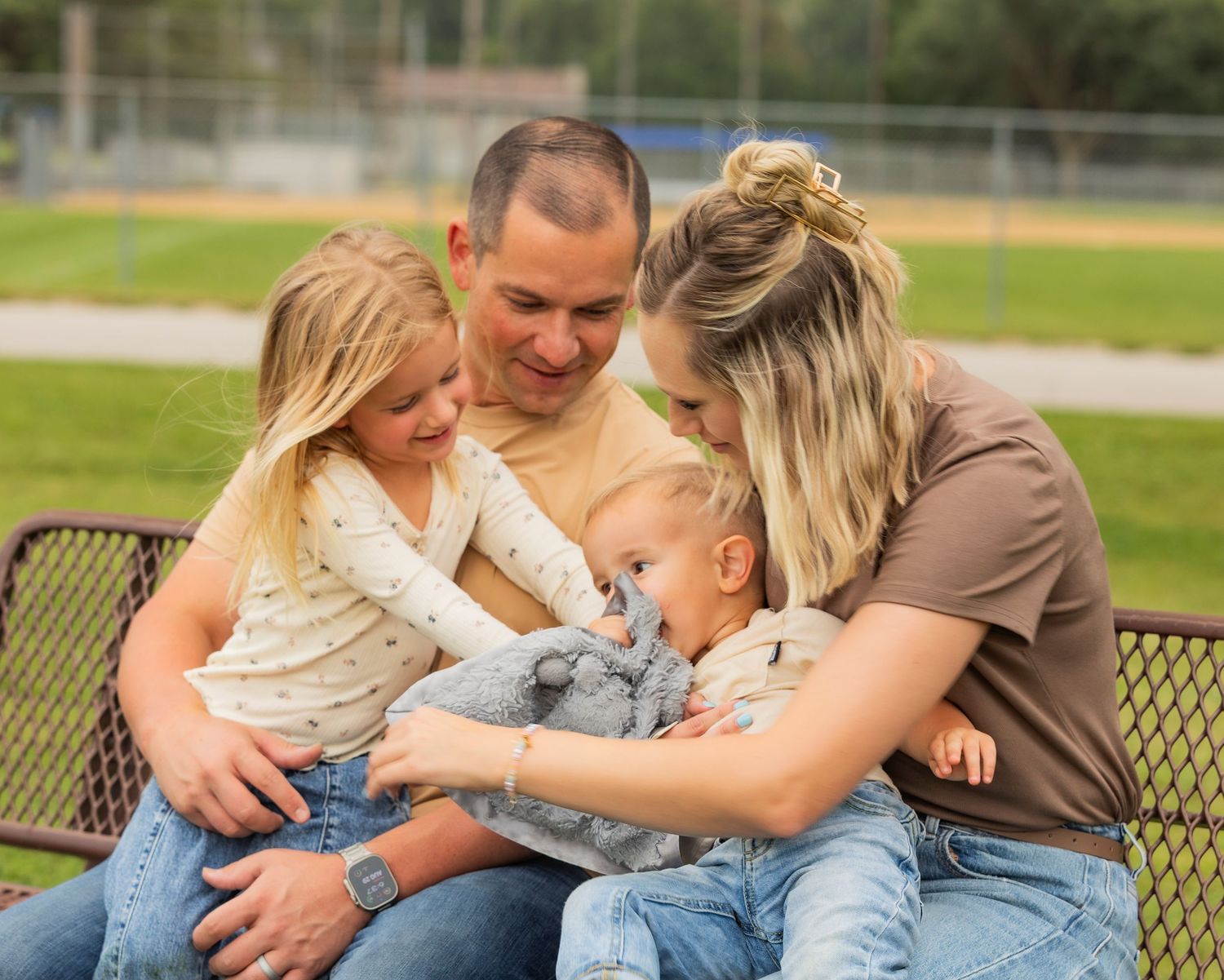 Family of four sitting on a bench outdoors, parents smiling at their baby and young blonde daughter.