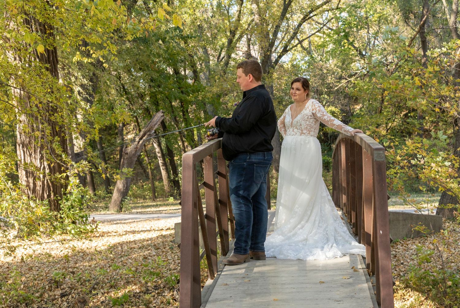 Wedding couple walking across a rustic wooden bridge surrounded by lush autumn foliage in a park setting.