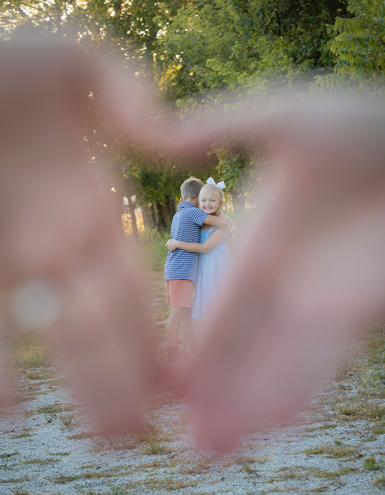 Two children hugging outdoors framed by heart-shaped hands in foreground, surrounded by lush green trees in warm sunlight.