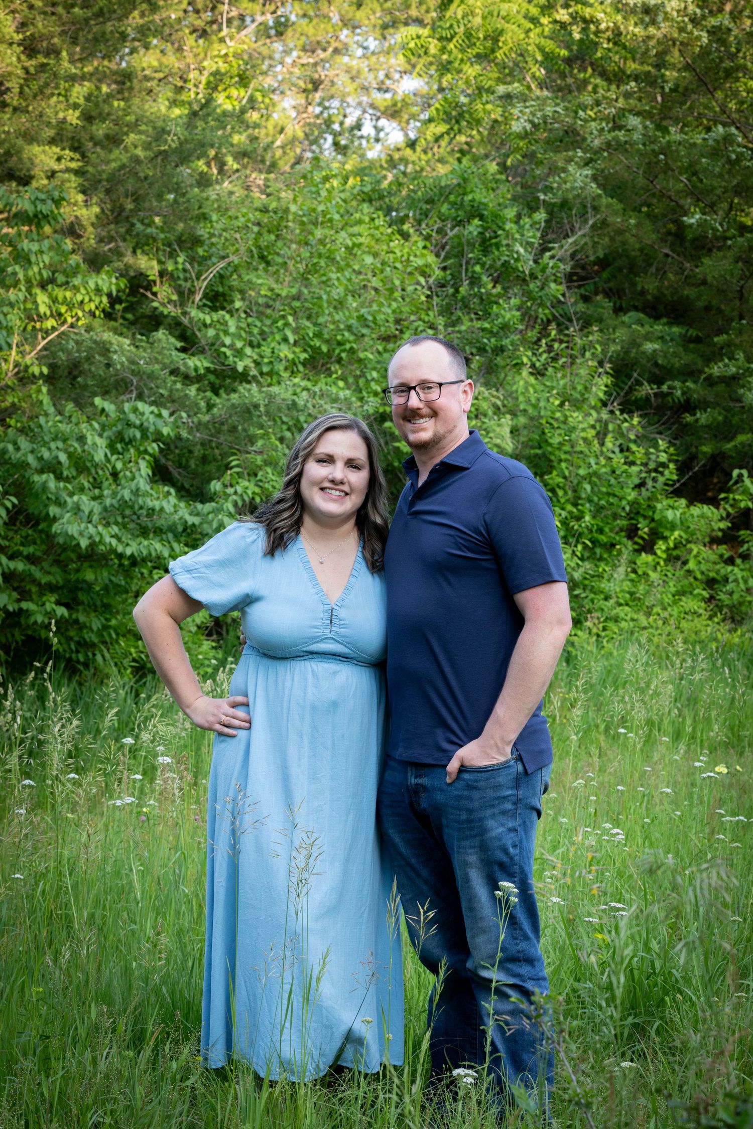 A couple poses together in a lush green meadow on a sunny summer day wearing blue and navy outfits.